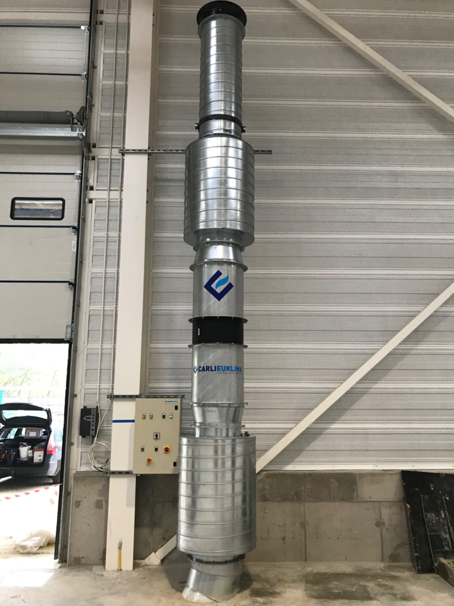 Metalowa kolumna wentylacyjna z logo firmy CARLIEUKLIMA, zainstalowana w hali przemysłowej z otwartą bramą i widocznym samochodem z narzędziami.
