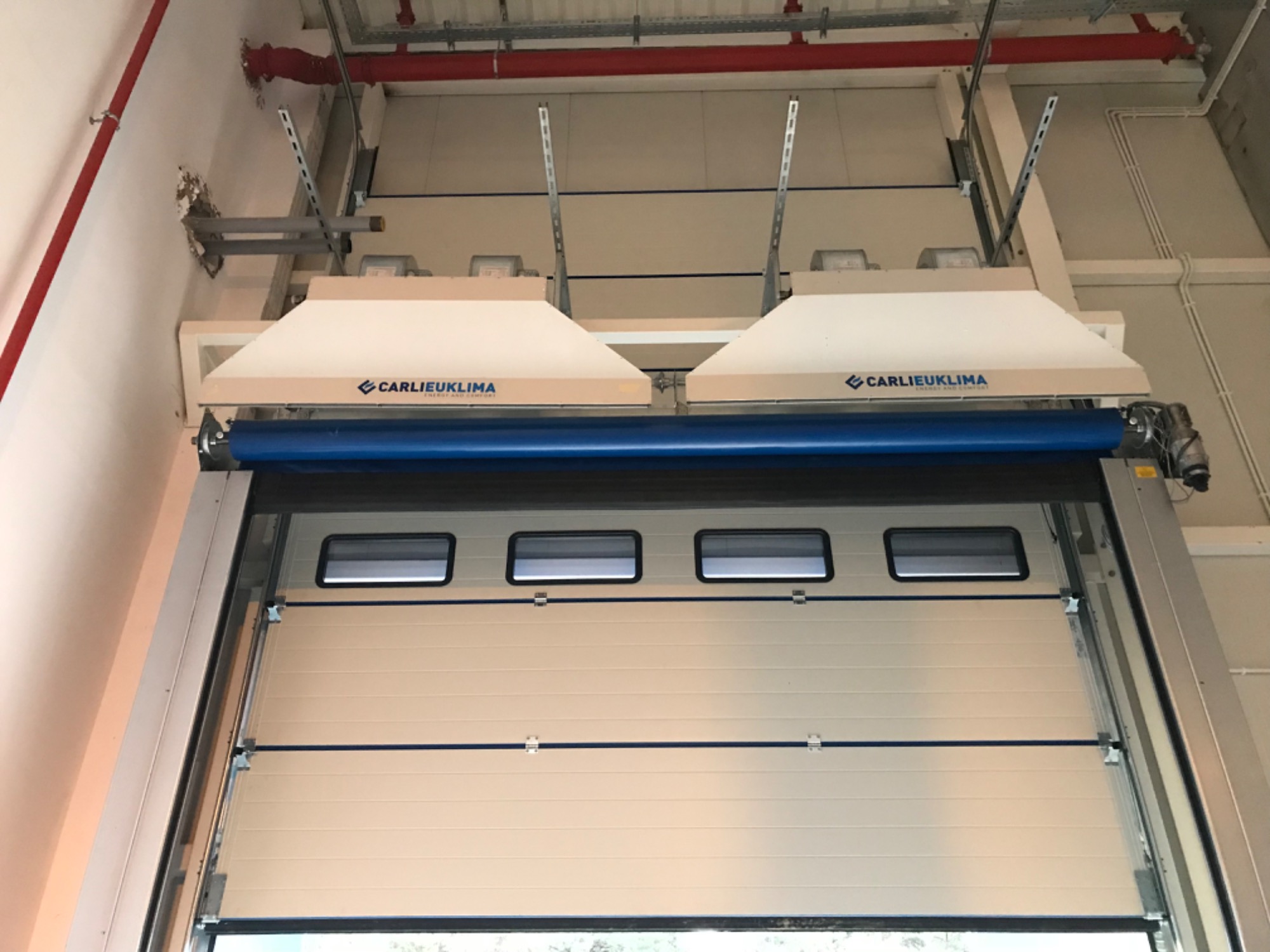 Otwarta segmentowa brama przemysłowa z zainstalowanymi dwoma nawiewami powietrza marki Carlieuklima, widoczne rury instalacji przeciwpożarowej pod sufitem.