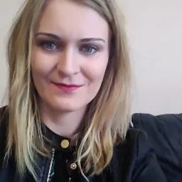 Portret kobiety o blond włosach i niebieskich oczach, z delikatnym makijażem ust i oczu, ubrana w czarną kurtkę z metalowymi guzikami.