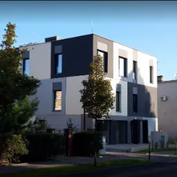 Nowoczesny, dwupiętrowy budynek biurowy z elewacją w odcieniach szarości i bieli, otoczony zielenią i niskim ogrodzeniem, widoczny z perspektywy ulicy w słoneczny dzień.