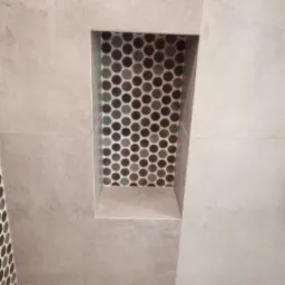 Wnęka w łazience wykończona szarą płytką imitującą beton i mozaiką z drobnych okrągłych płytek w odcieniach szarości i zieleni.