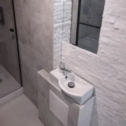 Nowoczesna łazienka z białą umywalką nablatową, szarą armaturą i mozaiką na ścianie. Widoczny fragment kabiny prysznicowej z czarnymi ramami.