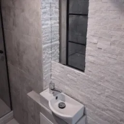 Nowoczesna łazienka z umywalką nablatową i lustrem w czarnej ramie. Ściany wyłożone płytkami imitującymi kamień, oświetlenie punktowe.