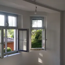 Pokój po gładziowaniu ścian i sufitu, z otwartymi oknami i butelką wody na podłodze.