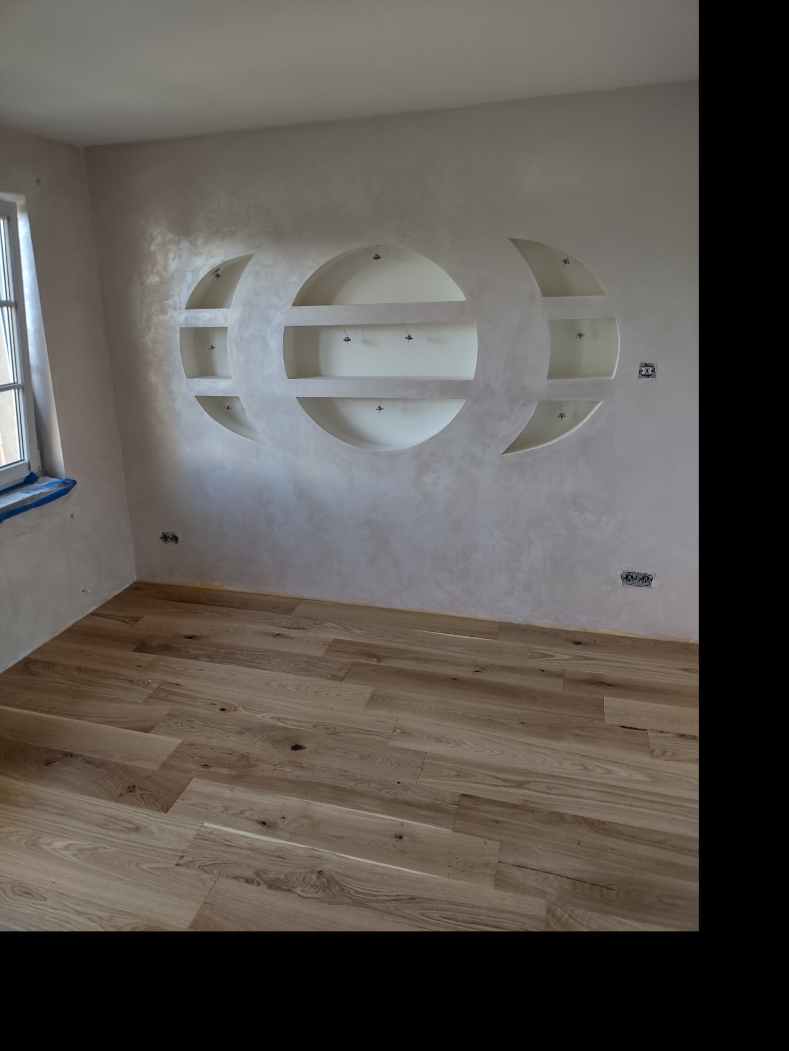 Jasne panele podłogowe i ściana z dekoracyjnymi wnękami w kształcie półksiężyca, oświetlonymi od góry, tworzącymi unikalny element wystroju wnętrza.