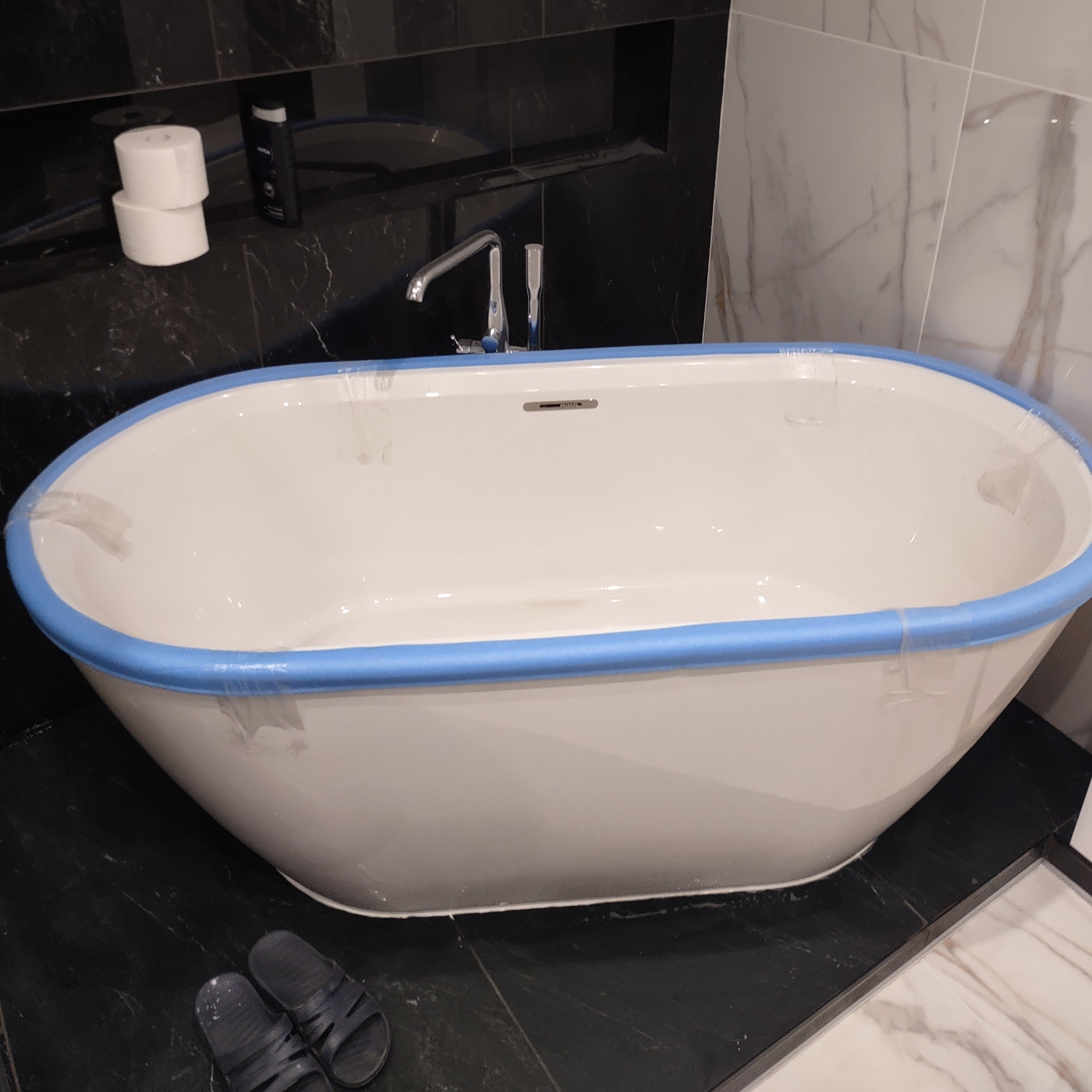 Nowoczesna, wolnostojąca wanna owalna z niebieskim zabezpieczeniem krawędzi, armatura, czarne płytki podłogowe i marmurowe ściany w trakcie remontu łazienki.