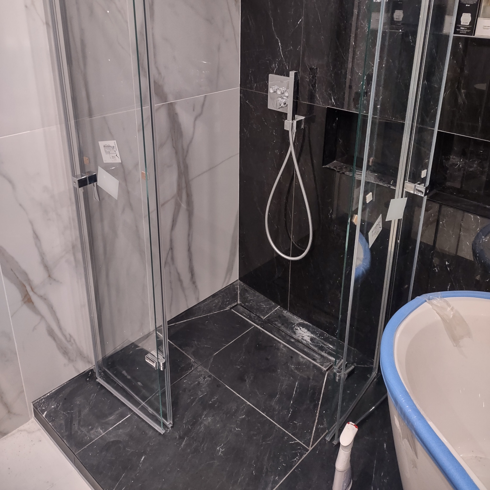 Nowoczesna kabina prysznicowa z czarno-białymi marmurowymi płytkami i chromowaną armaturą, częściowo widoczna wanna z niebieskim zabezpieczeniem.