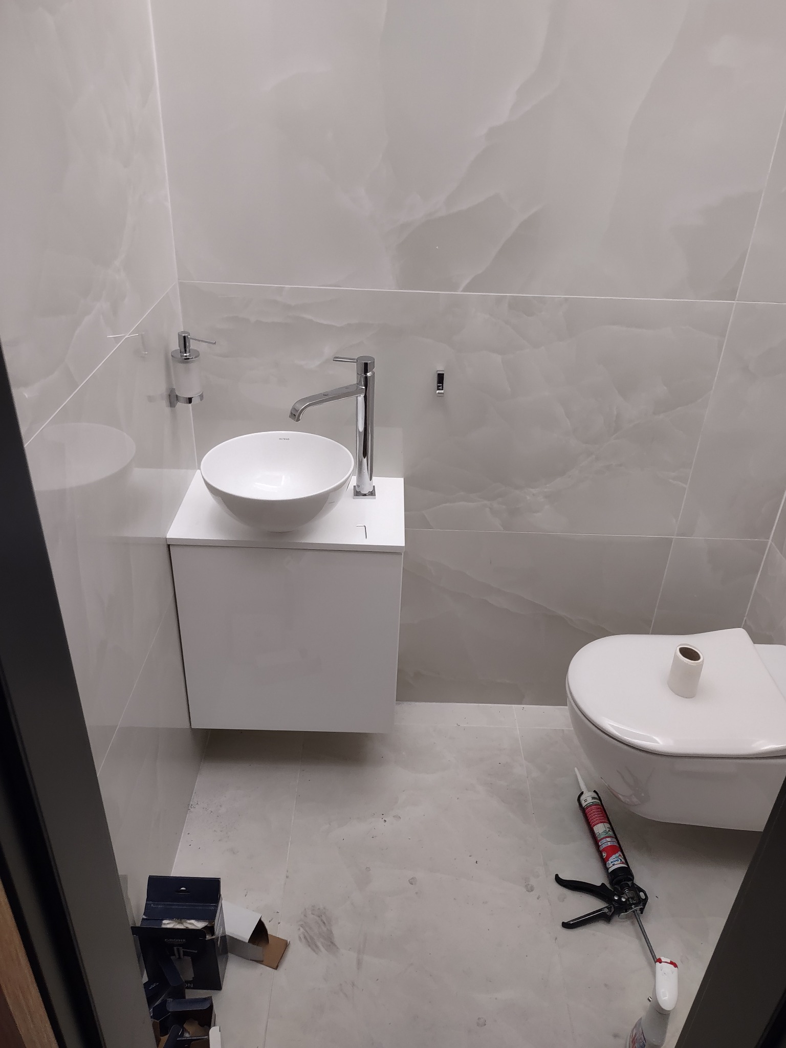 Nowoczesna, jasna łazienka po remoncie: okrągła umywalka nablatowa, wisząca toaleta, płytki imitujące marmur. Na podłodze narzędzia glazurnicze.