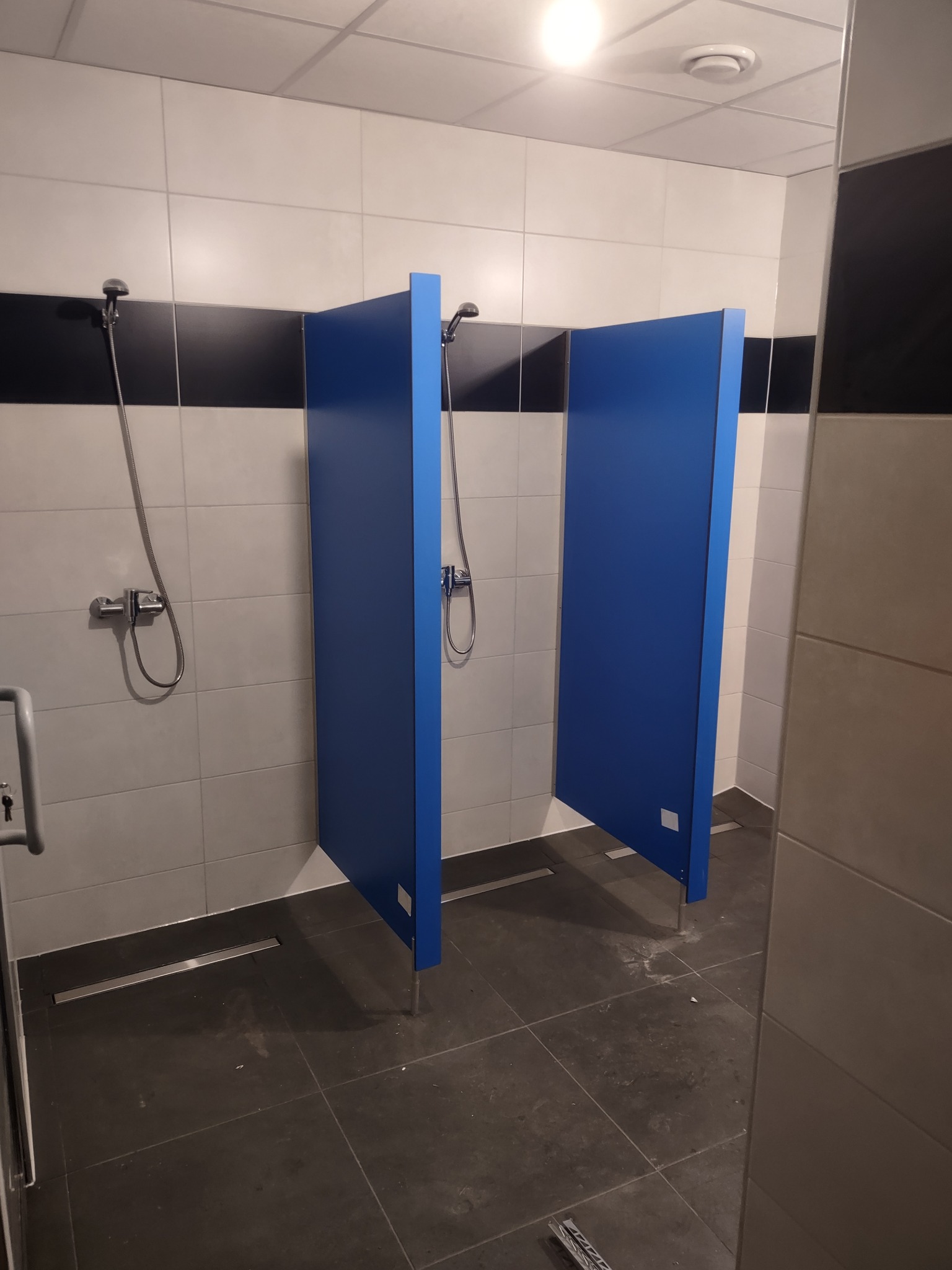 Wykończone kabiny prysznicowe z niebieskimi ściankami w publicznej łazience z szarymi i białymi płytkami. Widoczne baterie i odpływy liniowe.
