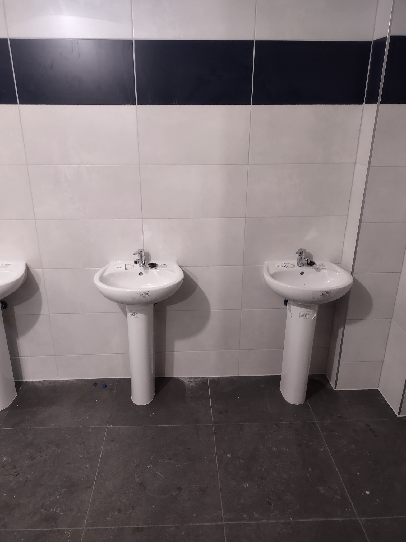 Wykończona łazienka z trzema białymi umywalkami na postumentach, szare płytki podłogowe i biało-czarne płytki ścienne. Widok z przodu, neutralne oświetlenie.