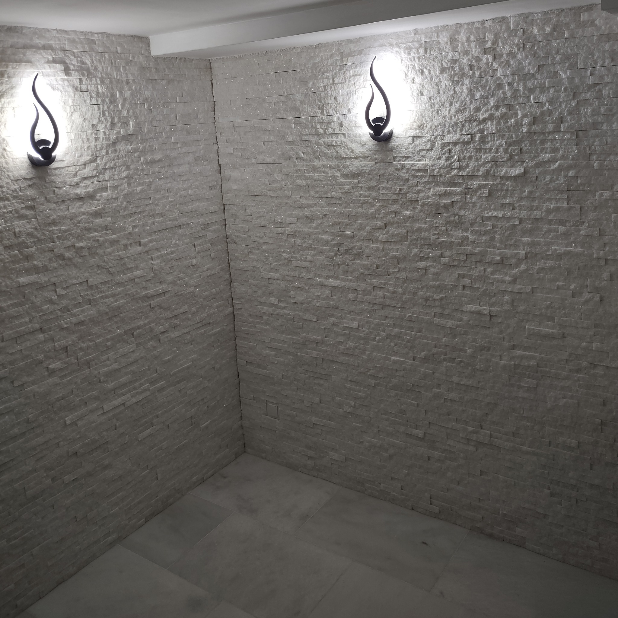 Biała łazienka wykończona dekoracyjną cegłą, oświetlona dwoma nowoczesnymi kinkietami w kształcie płomienia. Minimalistyczny styl, jasne wnętrze.