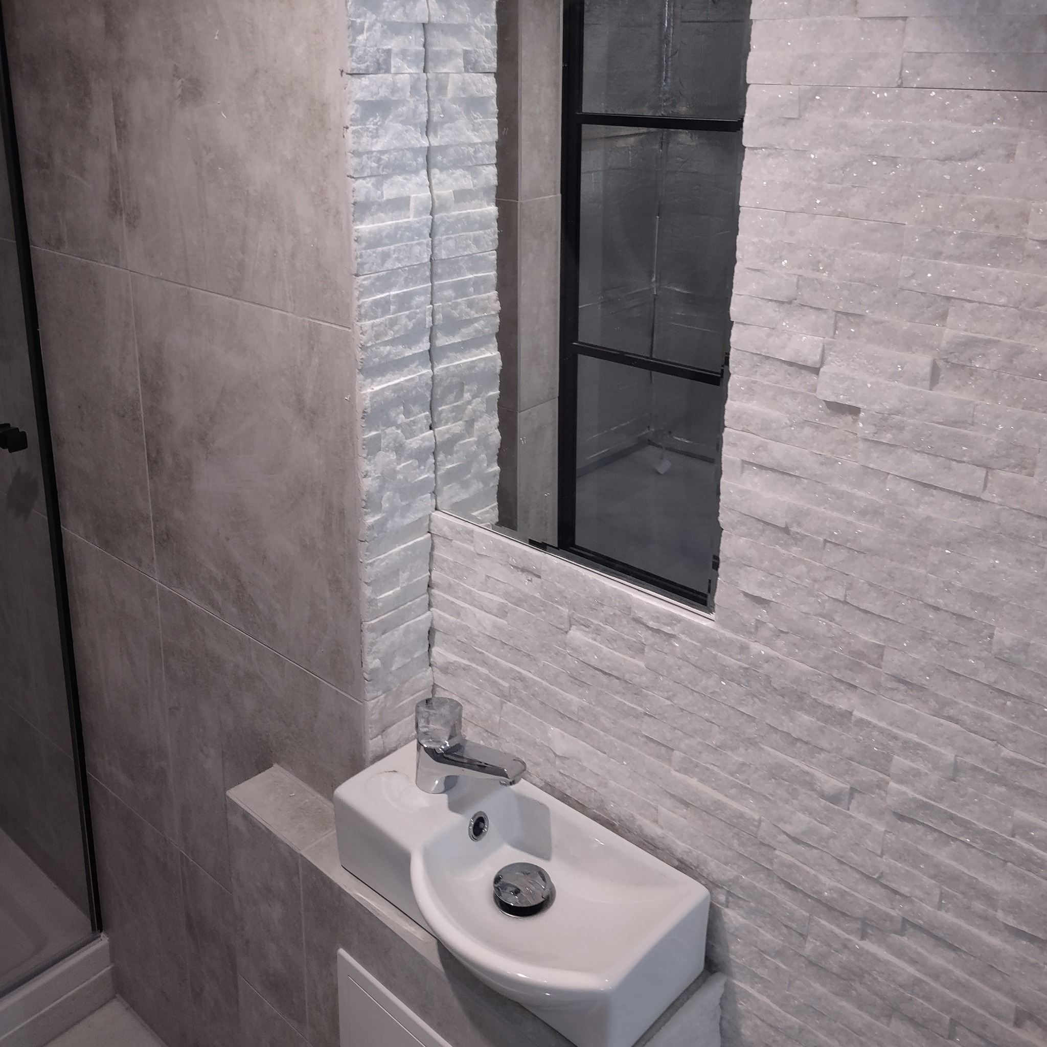 Nowoczesna łazienka z umywalką nablatową i lustrem w czarnej ramie. Ściany wyłożone płytkami imitującymi kamień, oświetlenie punktowe.