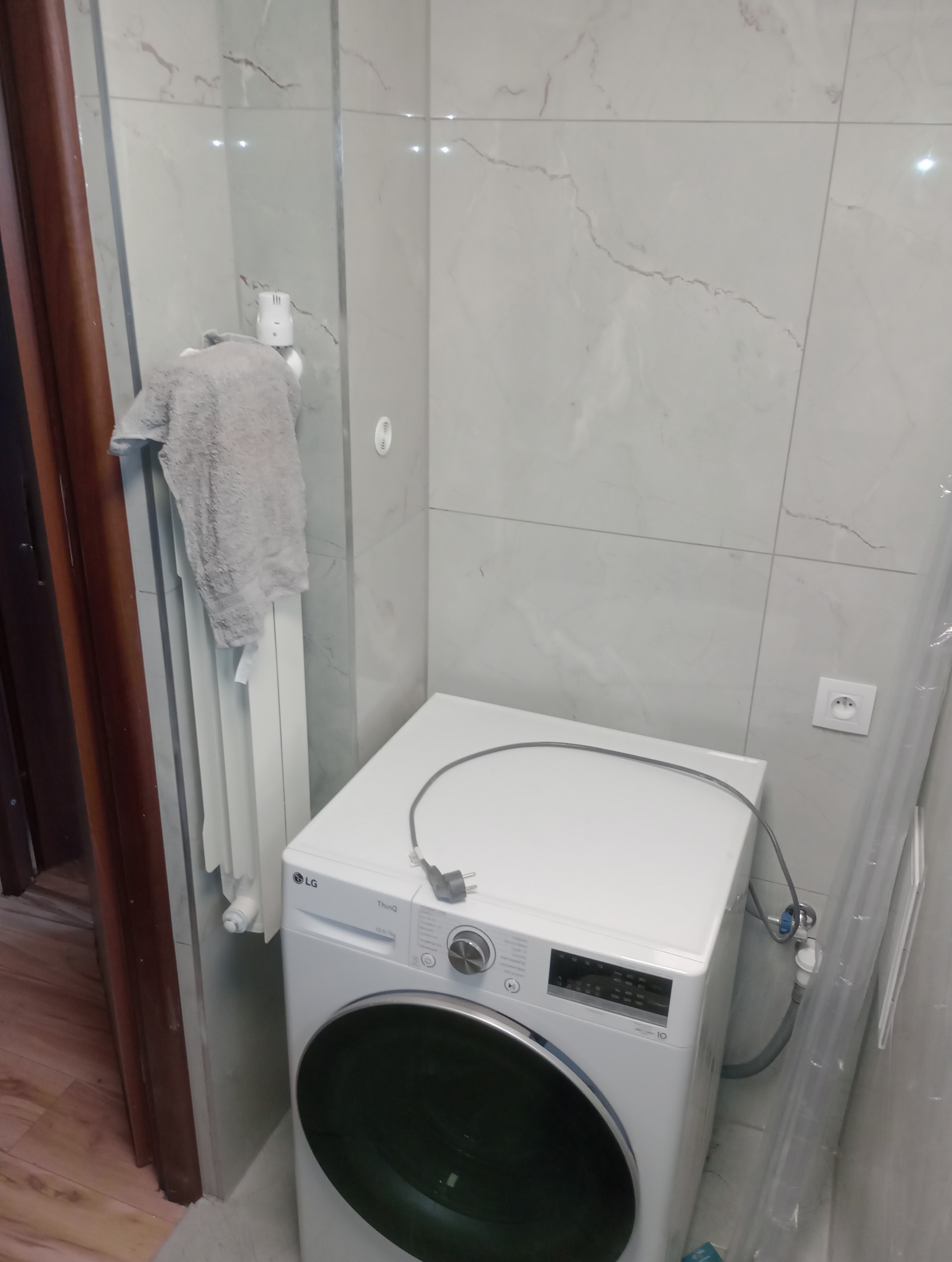 Jasna łazienka po remoncie: białe płytki imitujące marmur, grzejnik z ręcznikiem i nowa pralka LG. Widoczne przyłącza pralki.