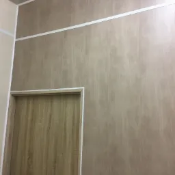 Ściana wykończona panelami imitującymi drewno z widoczną białą listwą dekoracyjną i ościeżnicą drewnianych drzwi z klamką.