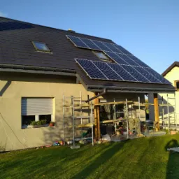 Dom jednorodzinny z zamontowanymi panelami słonecznymi na dachu, widoczne rusztowania i okna dachowe.