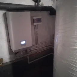 Biała jednostka wewnętrzna z wyświetlaczem, prawdopodobnie kotłownia, widoczne rury i zawory, obok zbiornik owinięty folią, surowe wykończenie ścian.