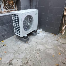 Nindo Nowoczesne Instalacje Domowe Przemysław Schimmelpfennig - Biała jednostka zewnętrzna klimatyzacji Midea, zamontowana na betonowych blokach przy ciemnej, kafelkowanej ścianie budynku. Widoczne liście na ziemi.