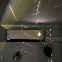 Nindo Nowoczesne Instalacje Domowe Przemysław Schimmelpfennig - Zamontowany klimatyzator marki Gree na ścianie, oświetlony dekoracyjnymi promieniami światła. Widoczny fragment drugiego klimatyzatora i element dekoracyjny na ścianie.