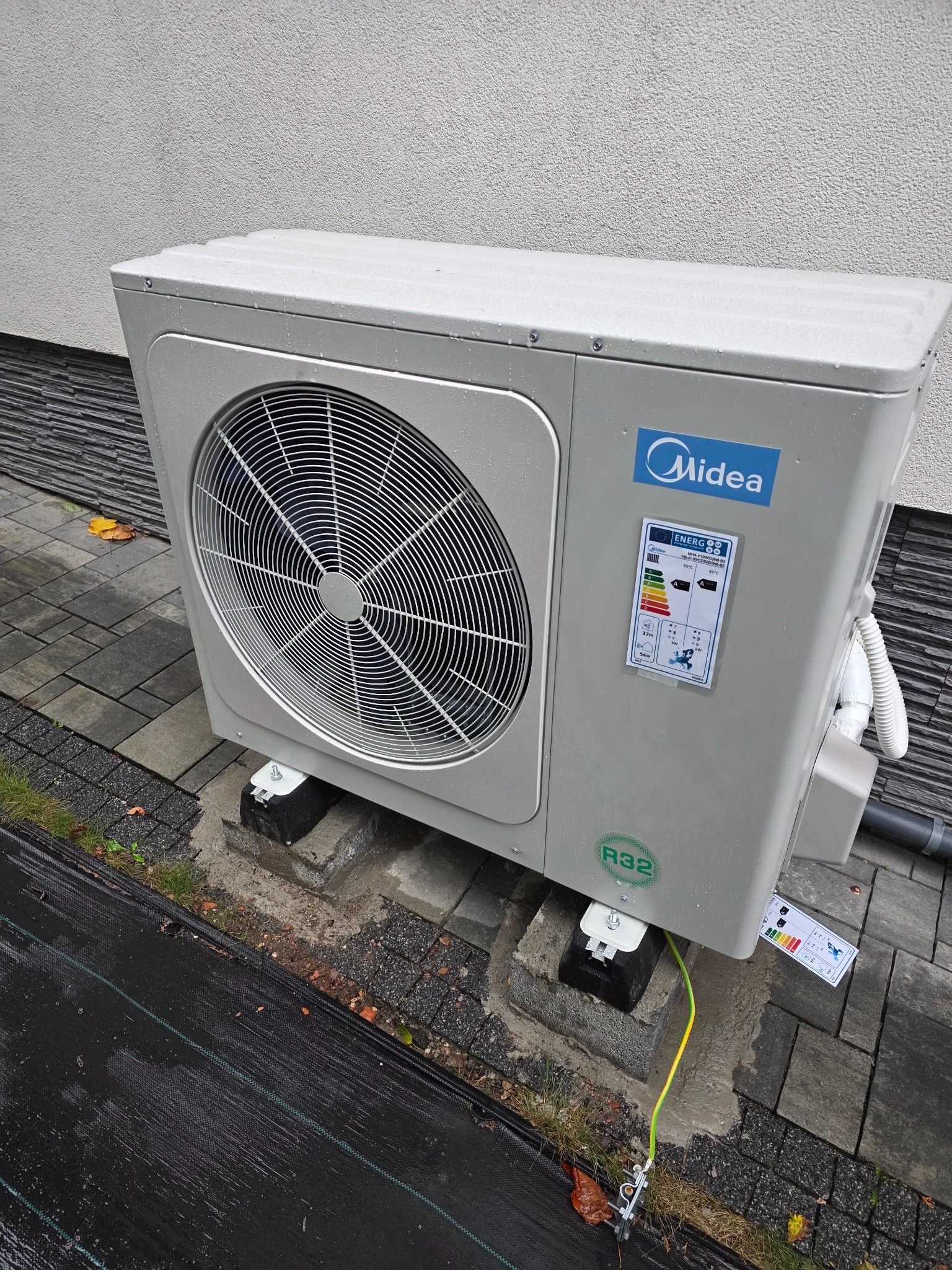 Zewnętrzna jednostka klimatyzacji Midea, zamontowana na betonowych podstawach, z widocznym uziemieniem i etykietą energetyczną, na tle kostki brukowej i elewacji budynku.