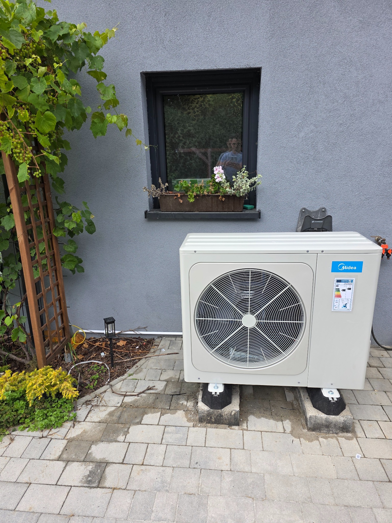 Biała jednostka zewnętrzna pompy ciepła Midea, umieszczona na betonowych podstawach, na tle szarej ściany z oknem i zielenią. Widoczne elementy instalacji.