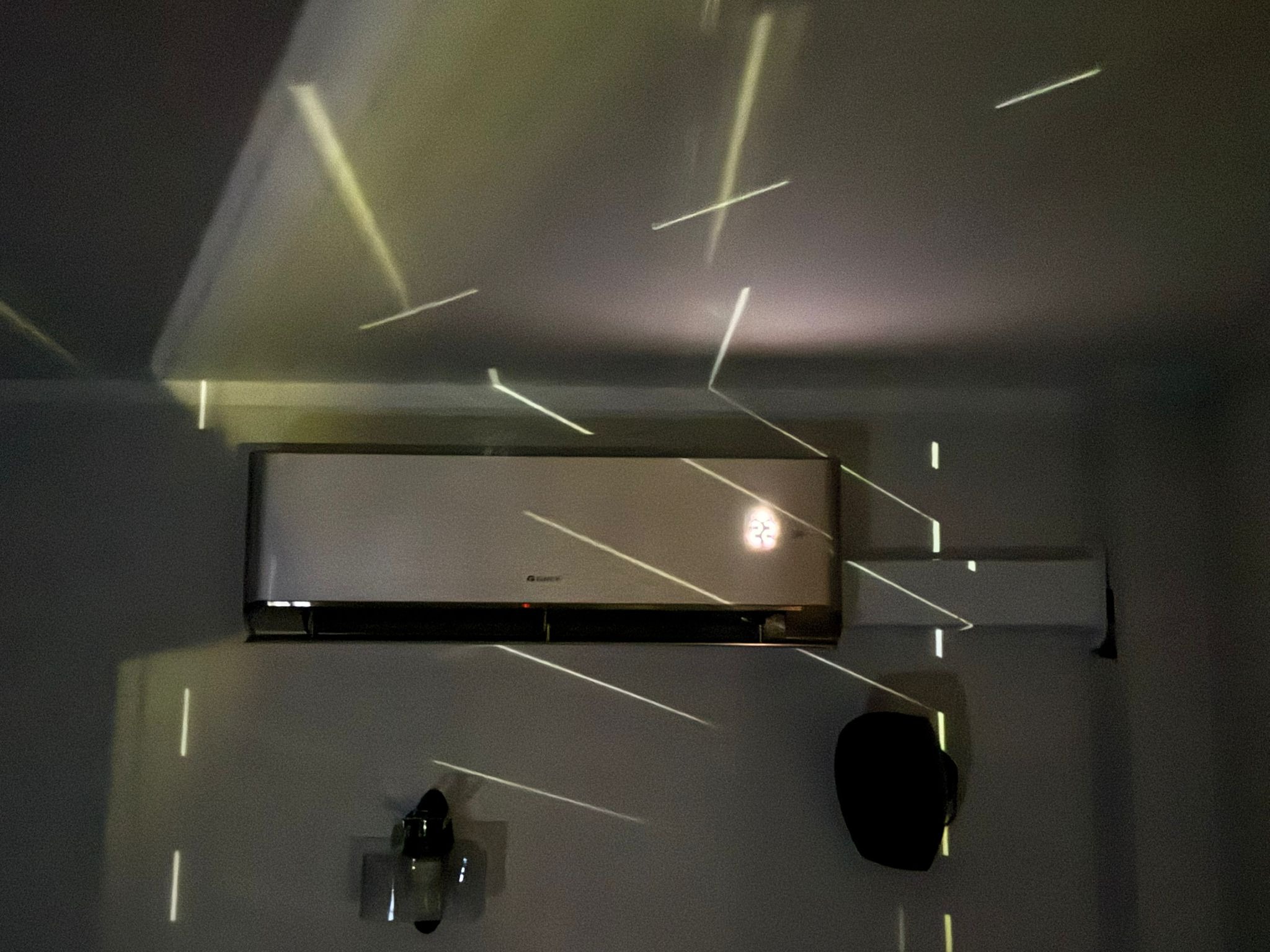 Zamontowany klimatyzator marki Gree na ścianie, oświetlony dekoracyjnymi promieniami światła. Widoczny fragment drugiego klimatyzatora i element dekoracyjny na ścianie.