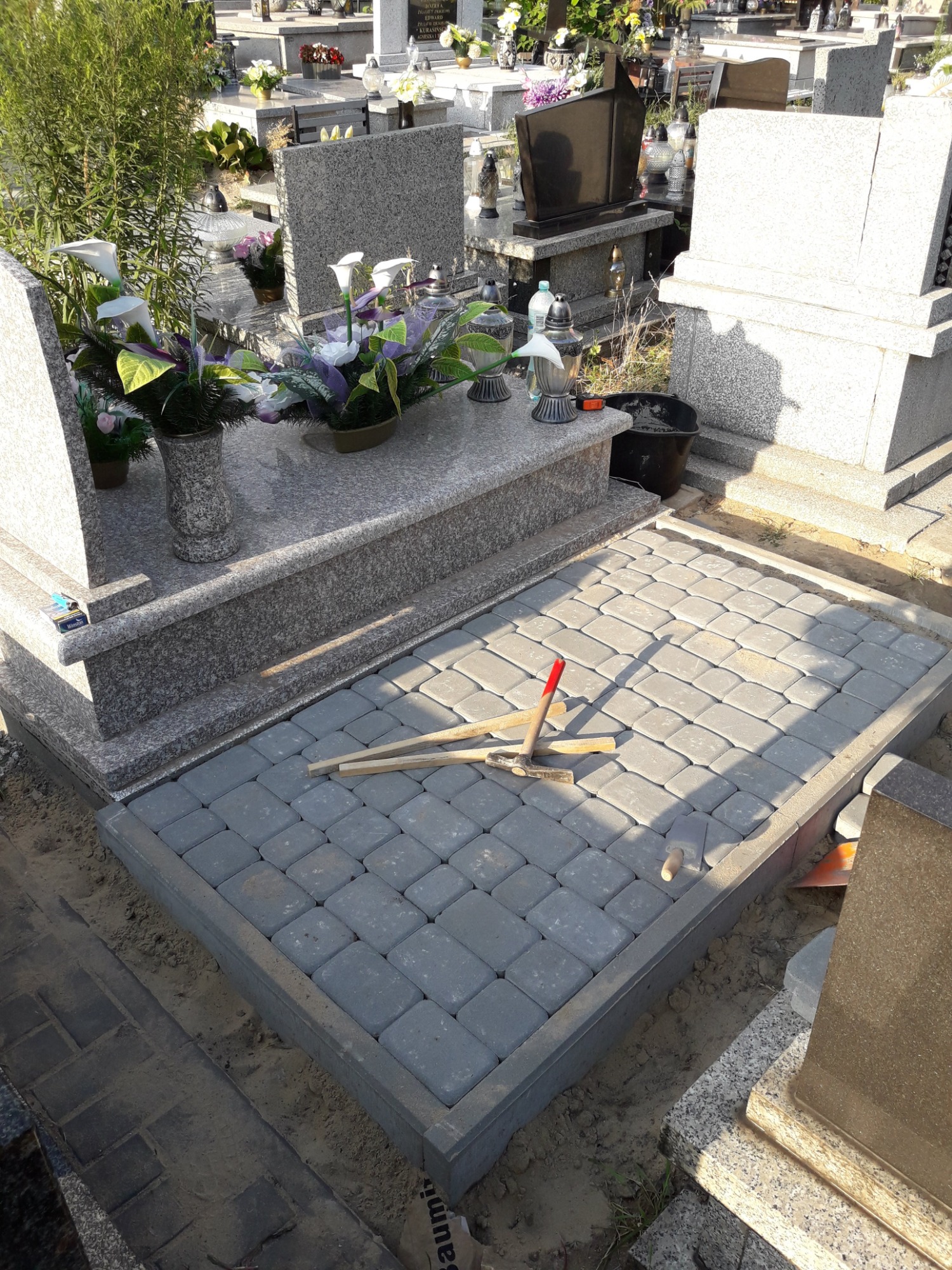 Częściowo wyłożona kostką przestrzeń przed nagrobkiem z narzędziami do układania kostki brukową, widoczne inne nagrobki w tle.