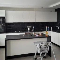 Nowoczesna kuchnia z białymi szafkami i czarną glazurą na ścianie, wyspa kuchenna z czarnym blatem, widoczne narzędzia i akcesoria na blacie, w tle zabudowa kuchenna z piekarnikiem i płytą grzewczą...