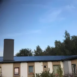 Dwukondygnacyjny budynek z niebieskim dachem i dużym, szarym kominem na tle błękitnego nieba i zieleni drzew.