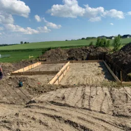 Przygotowany teren pod budowę domu z drewnianym szalunkiem fundamentów, widoczny pracownik i narzędzia na tle zielonych pól i błękitnego nieba.