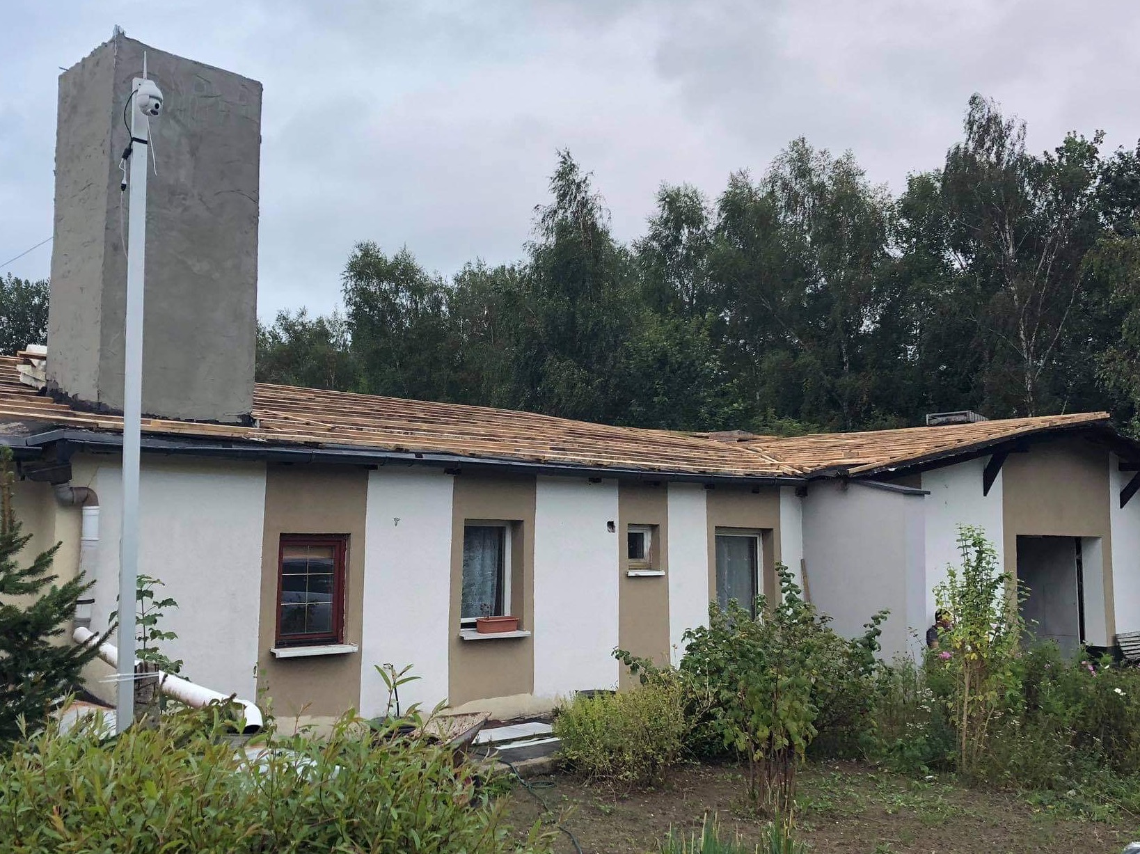 Budynek jednorodzinny z częściowo zdjętym dachem, widoczne drewniane łaty przygotowane pod nowe pokrycie dachowe, obok komina zamontowana kamera monitoringu.