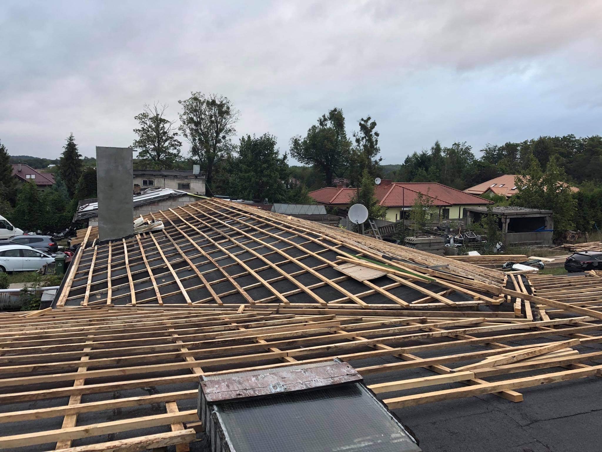 Konstrukcja drewniana dachu w trakcie budowy, widoczne deski tworzące szkielet pod przyszłe pokrycie, w tle zabudowania mieszkalne i zieleń.