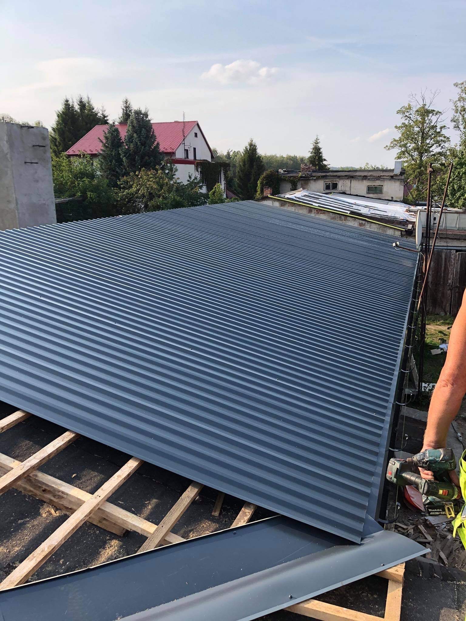 Montaż blachy trapezowej na dachu budynku, widoczne drewniane krokwie i wkrętarka w dłoni montera, w tle zabudowa jednorodzinna.