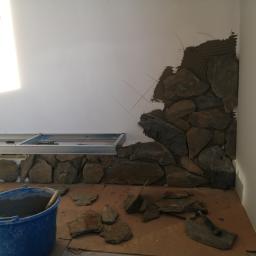 Remonty-Wykonczenia Sobieski - Fragment ściany w trakcie wykańczania kamieniem dekoracyjnym, widoczny metalowy stelaż pod sufitem, wiadro z zaprawą i narzędzia.