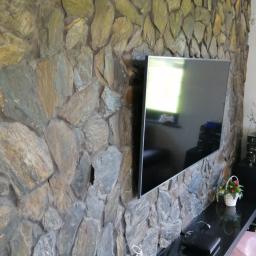 Remonty-Wykonczenia Sobieski - Ściana wykończona kamieniem naturalnym w odcieniach szarości i beżu, z zamontowanym telewizorem i czarnym blatem pod nim, na którym stoją dekoracje.