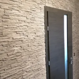 Ściana wykończona dekoracyjnym kamieniem w odcieniach beżu, z widocznymi poziomymi warstwami i fakturą, sąsiadująca z ciemnoszarą, nowoczesną stolarką drzwiową z pionowym przeszkleniem.