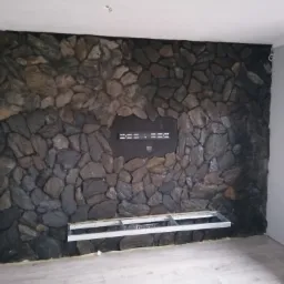 Ściana wykończona kamieniem dekoracyjnym, z przygotowanymi otworami na montaż telewizora i kominka elektrycznego, widoczne metalowe profile montażowe.