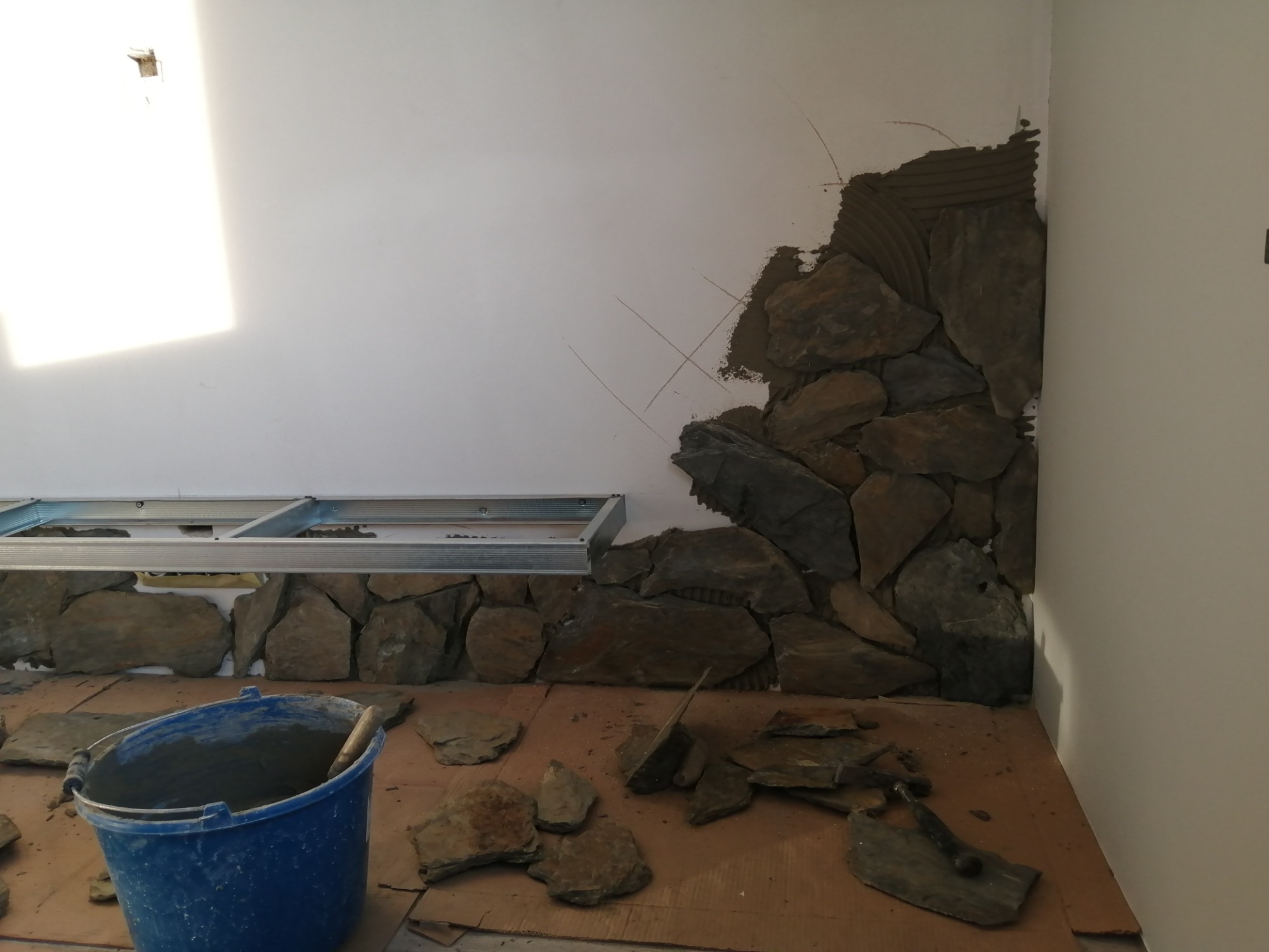 Fragment ściany w trakcie wykańczania kamieniem dekoracyjnym, widoczny metalowy stelaż pod sufitem, wiadro z zaprawą i narzędzia.