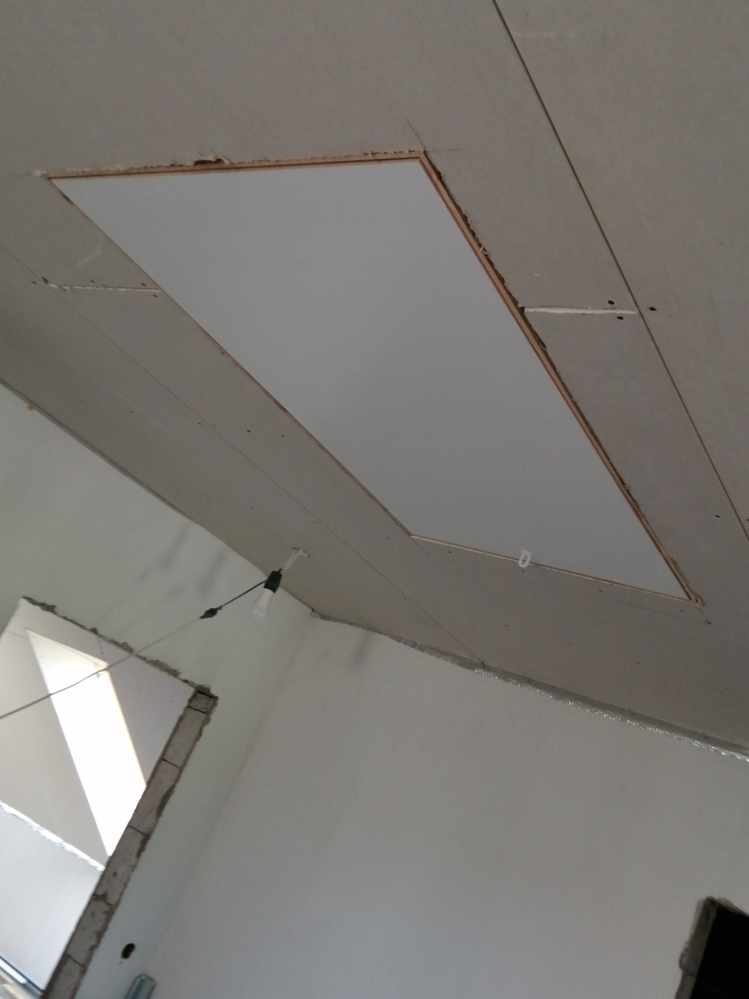 Montaż płyty gipsowo-kartonowej w suficie, widoczne niedoskonałości wykończenia wokół otworu rewizyjnego, surowy stan wnętrza z wiszącą żarówką.