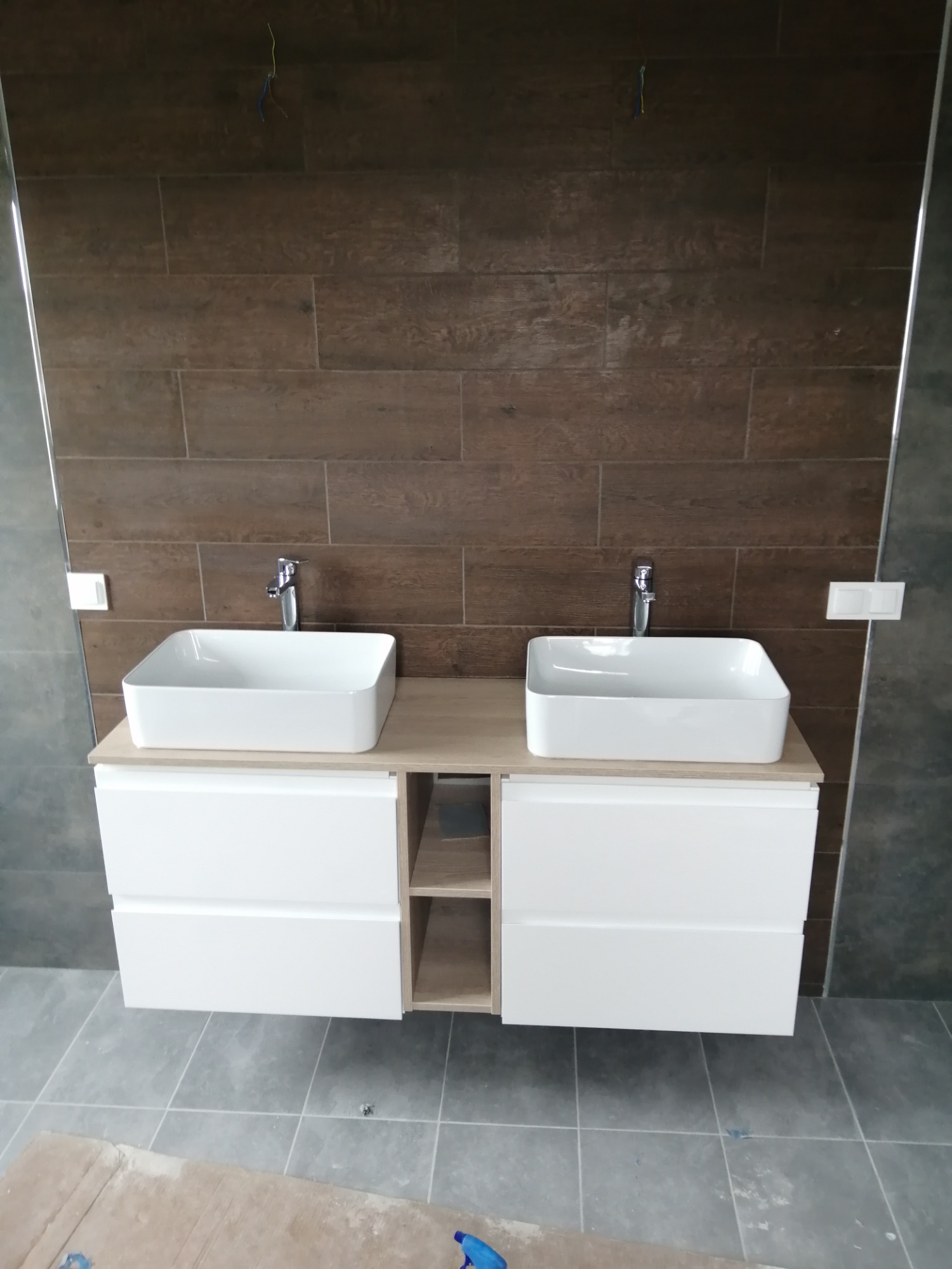 Nowoczesna łazienka z dwoma białymi umywalkami nablatowymi na szafce z szufladami i otwartą półką, brązowe płytki imitujące drewno na ścianie, szare płytki podłogowe, widoczne kable wystające ze...