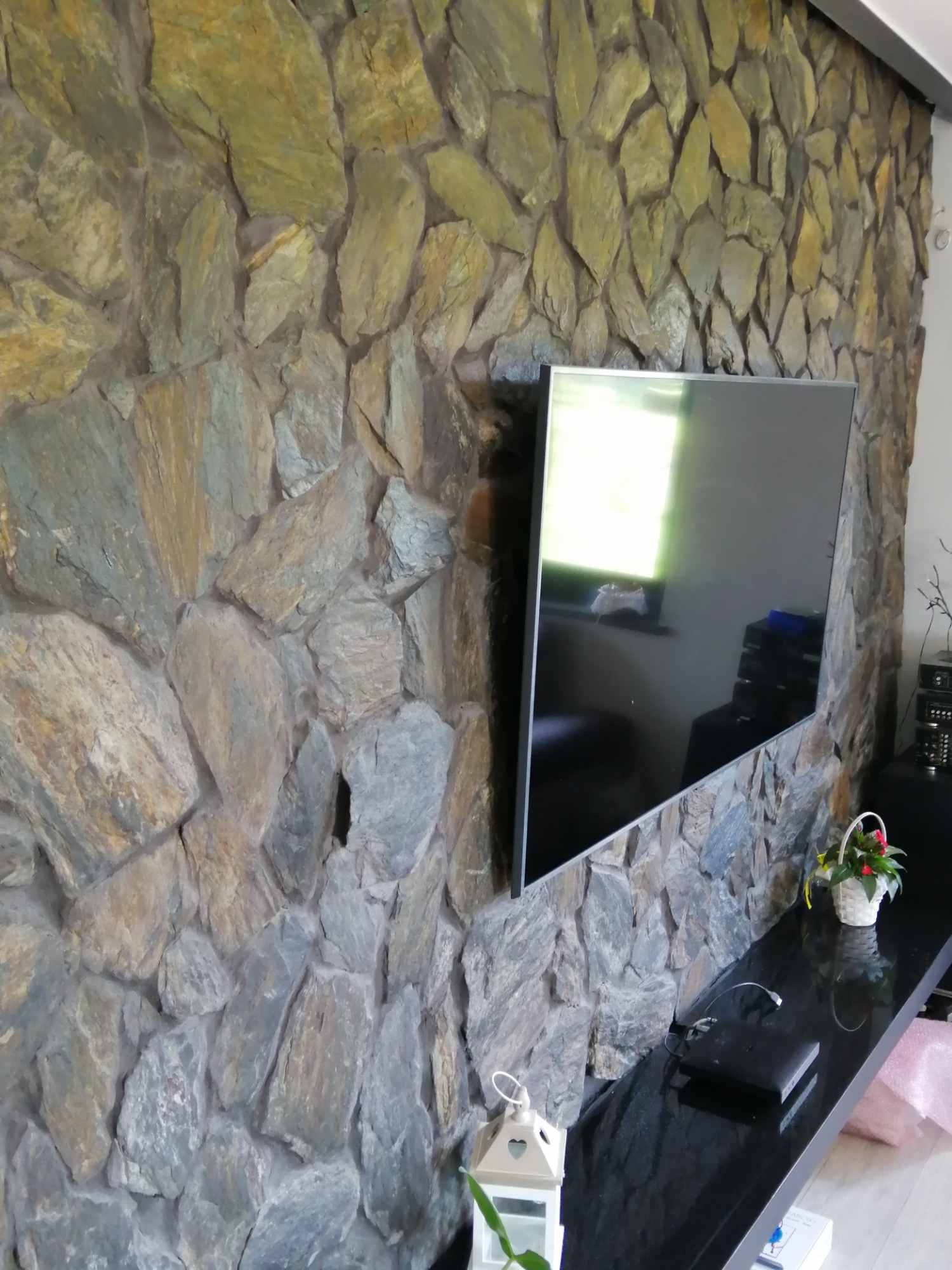 Ściana wykończona kamieniem naturalnym w odcieniach szarości i beżu, z zamontowanym telewizorem i czarnym blatem pod nim, na którym stoją dekoracje.