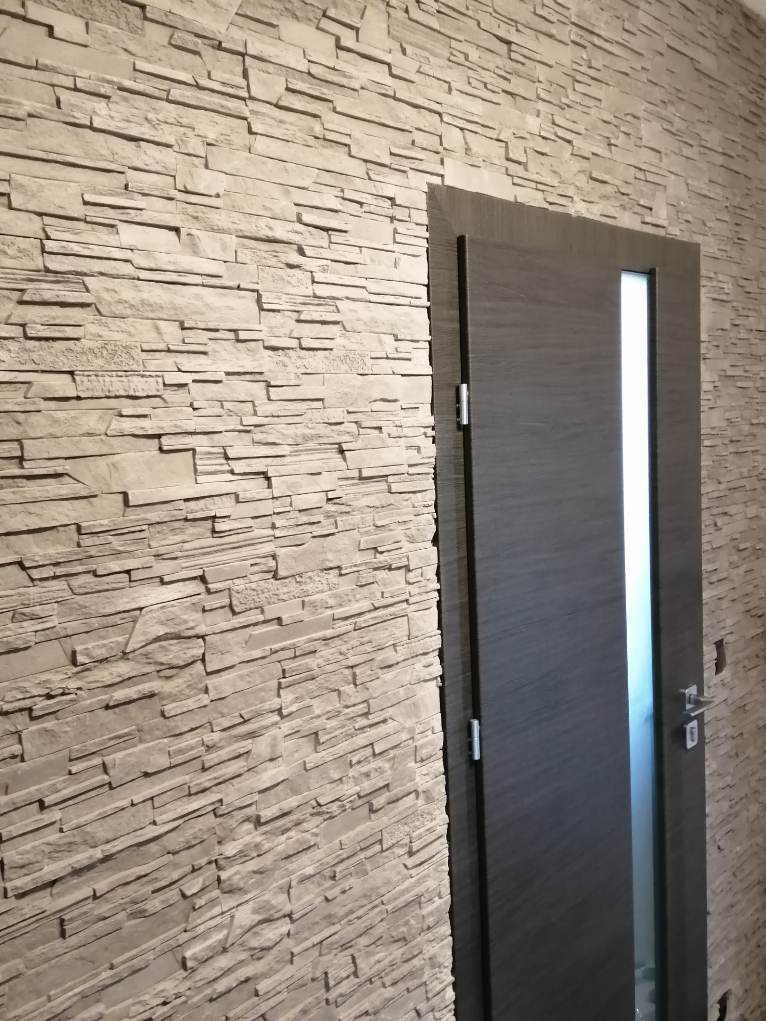 Ściana wykończona dekoracyjnym kamieniem w odcieniach beżu, z widocznymi poziomymi warstwami i fakturą, sąsiadująca z ciemnoszarą, nowoczesną stolarką drzwiową z pionowym przeszkleniem.