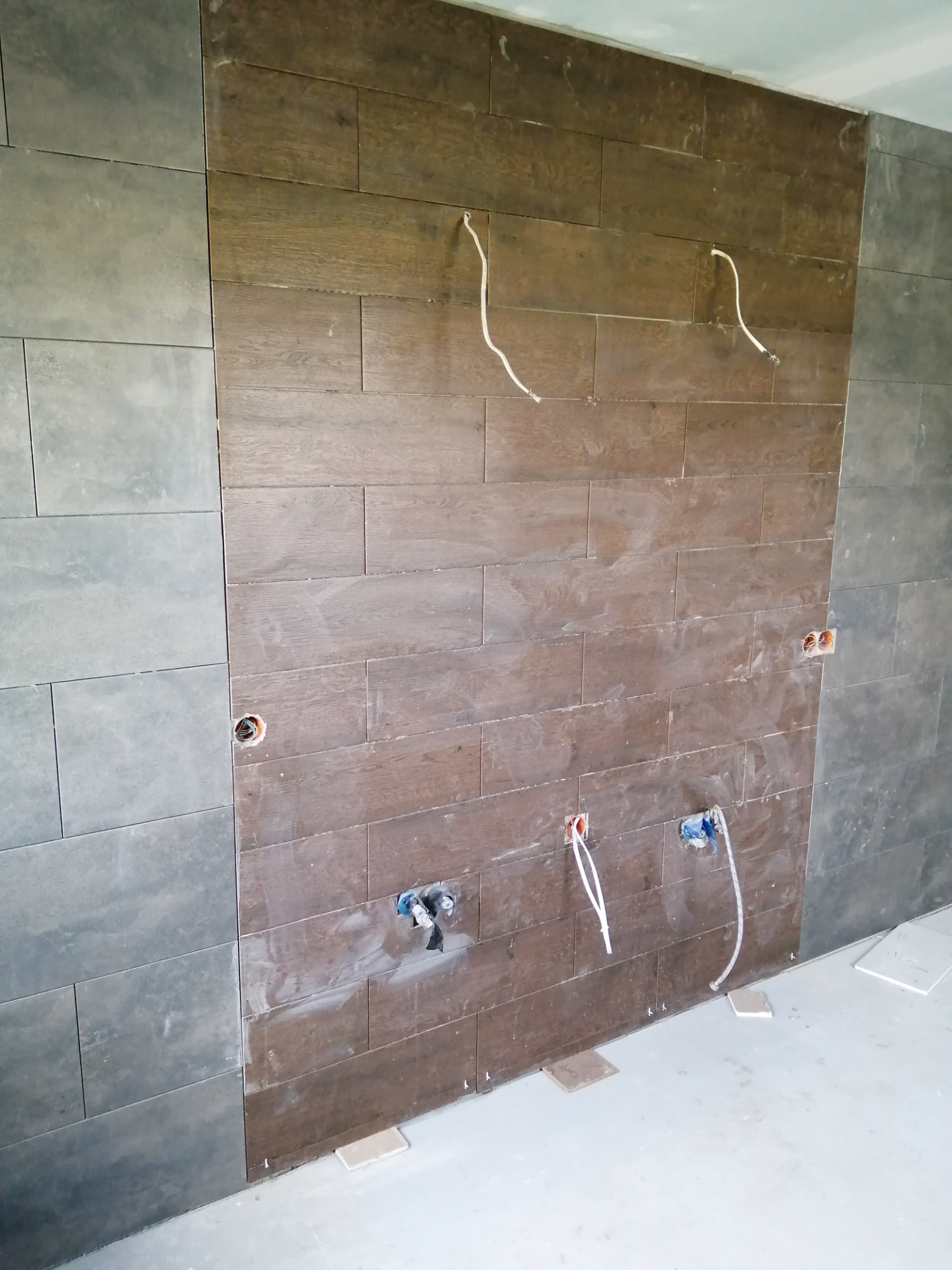 Ściana w trakcie wykańczania z fragmentami szarych i drewnopodobnych płytek, widoczne wystające kable i rury, oraz płytki dystansowe na podłodze.