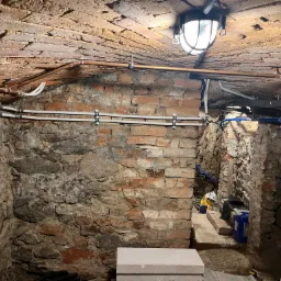 Piwnica z kamiennymi ścianami i ceglanym sklepieniem, widoczne rury instalacji, oświetlenie robocze i materiały budowlane na podłodze.
