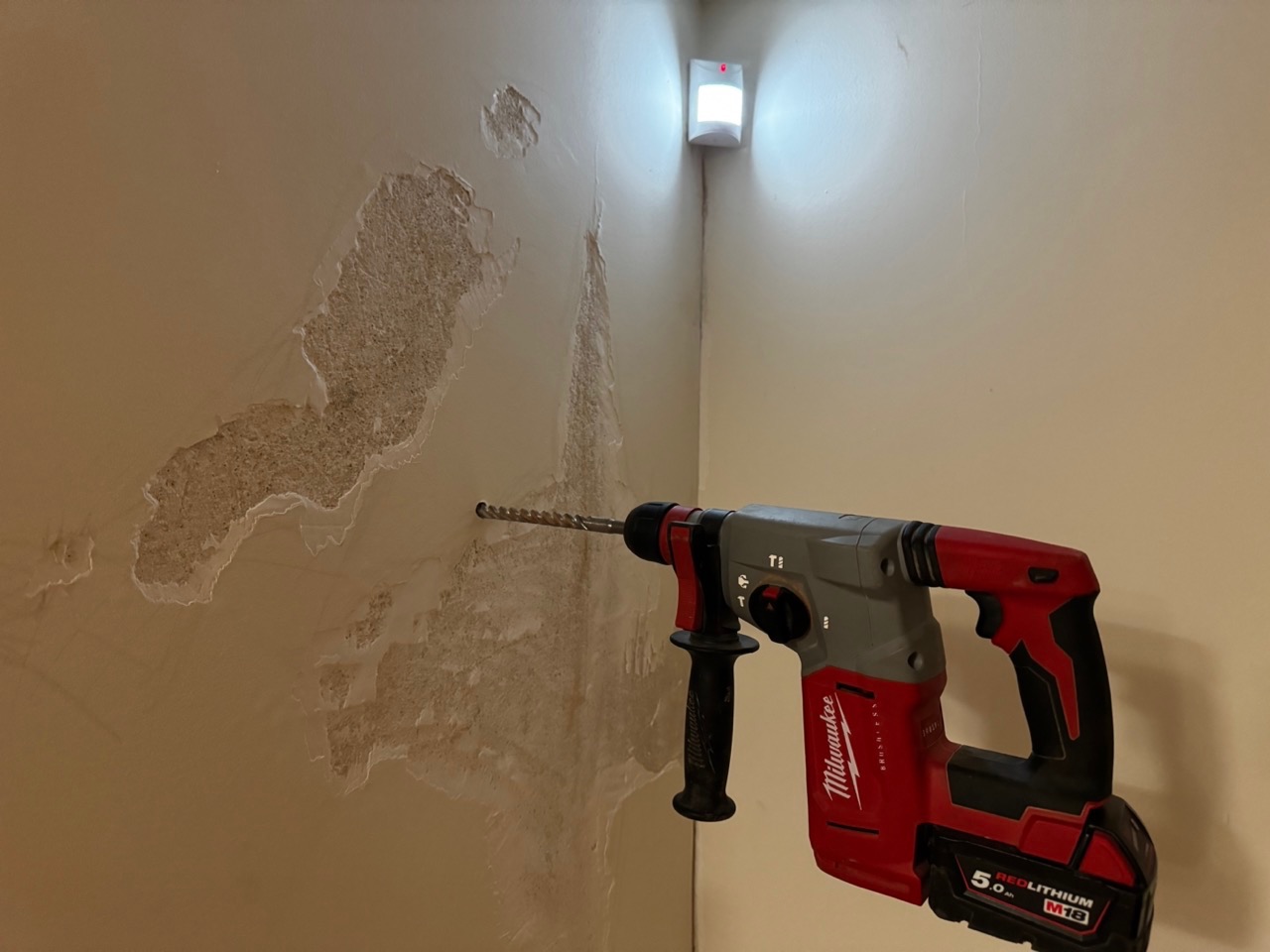 Wiertarka udarowa Milwaukee z wiertłem w ścianie z widocznymi śladami wilgoci i odpadającym tynkiem, oświetlona lampą z czujnikiem ruchu.