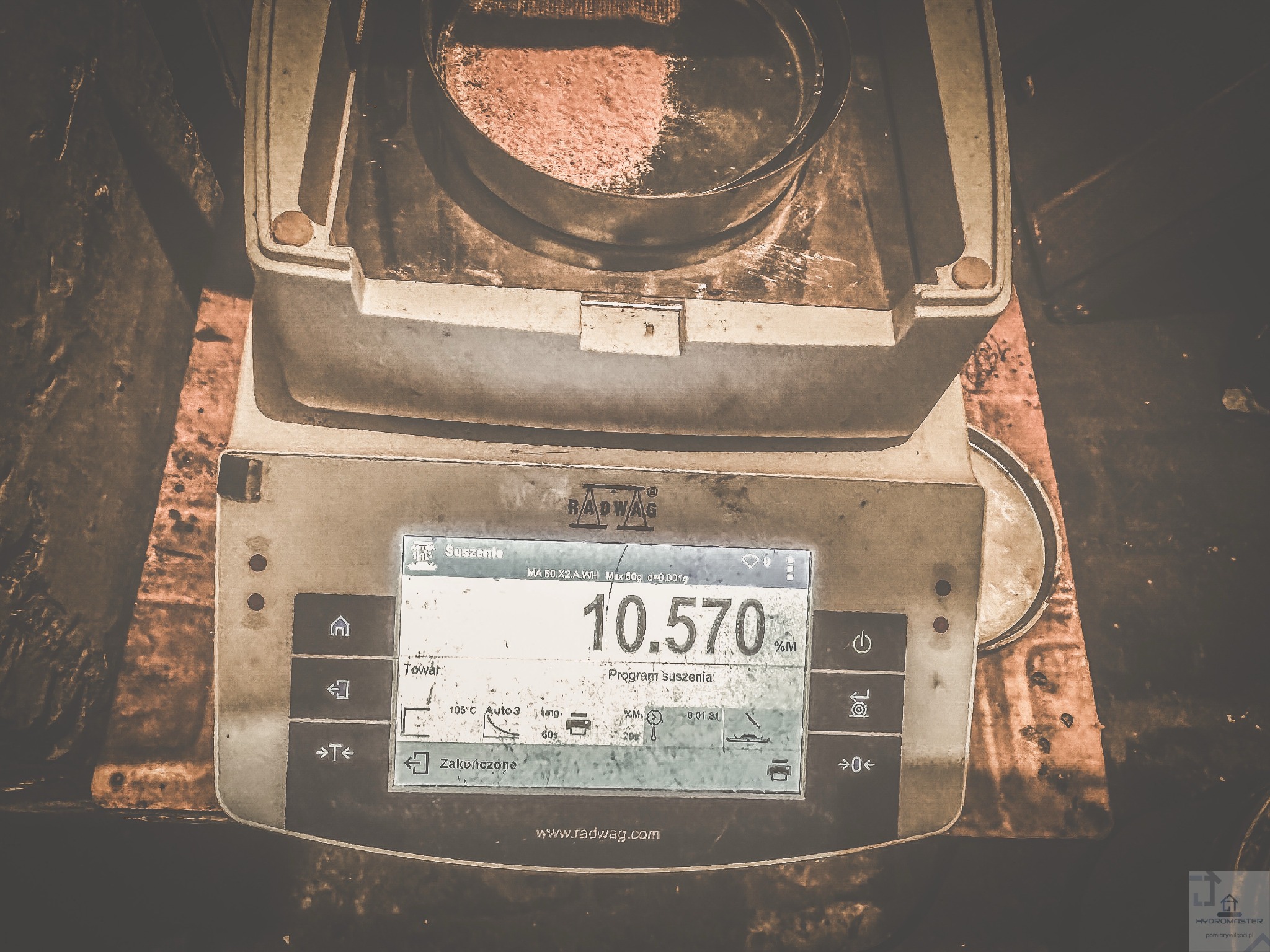 Waga laboratoryjna Radwag z próbką materiału w metalowym naczyniu, wyświetlająca wynik pomiaru wilgotności. Urządzenie używane do osuszania i badania materiałów budowlanych.