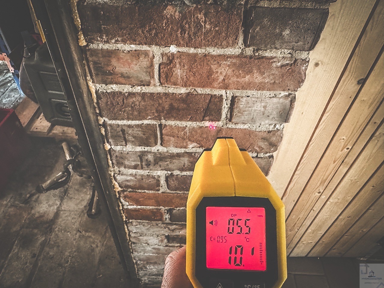 Pomiar wilgotności ściany z cegły za pomocą wilgotnościomierza elektronicznego z czerwonym wyświetlaczem, widoczne drewniane elementy konstrukcyjne.