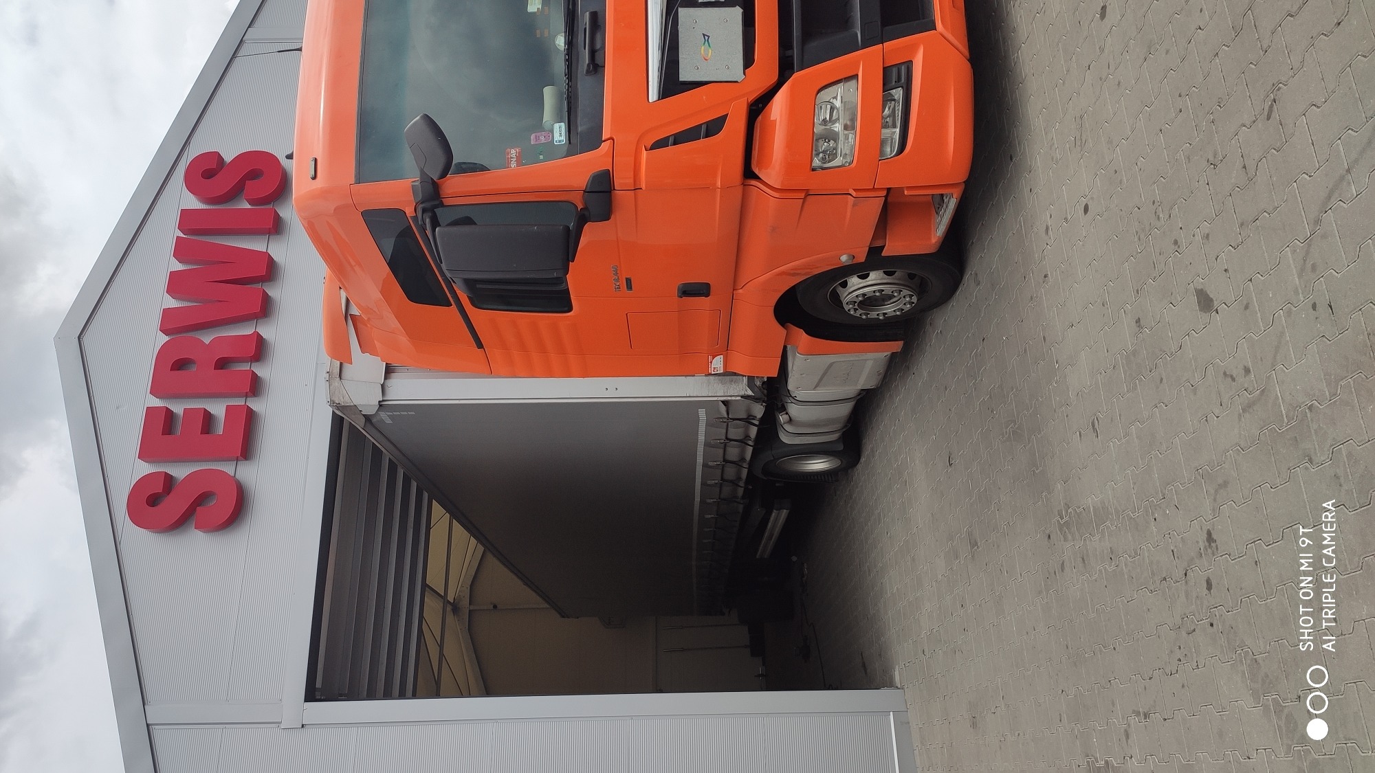 Pomarańczowa ciężarówka wjeżdża do hali serwisowej z dużym czerwonym napisem 'SERWIS' na elewacji budynku.