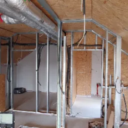 Wnętrze poddasza w trakcie adaptacji z widoczną konstrukcją ścian działowych z profili stalowych, przewodami elektrycznymi w peszlach i rurami wentylacyjnymi pod stropem, oświetlone lampą roboczą.