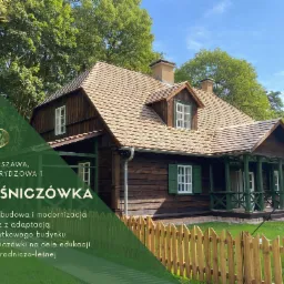 Odrestaurowana, drewniana leśniczówka z brązowymi ścianami, jasnobrązowym dachem z gontu, białymi ramami okiennymi i zielonymi okiennicami, otoczona zielenią drzew i niskim, drewnianym płotem.