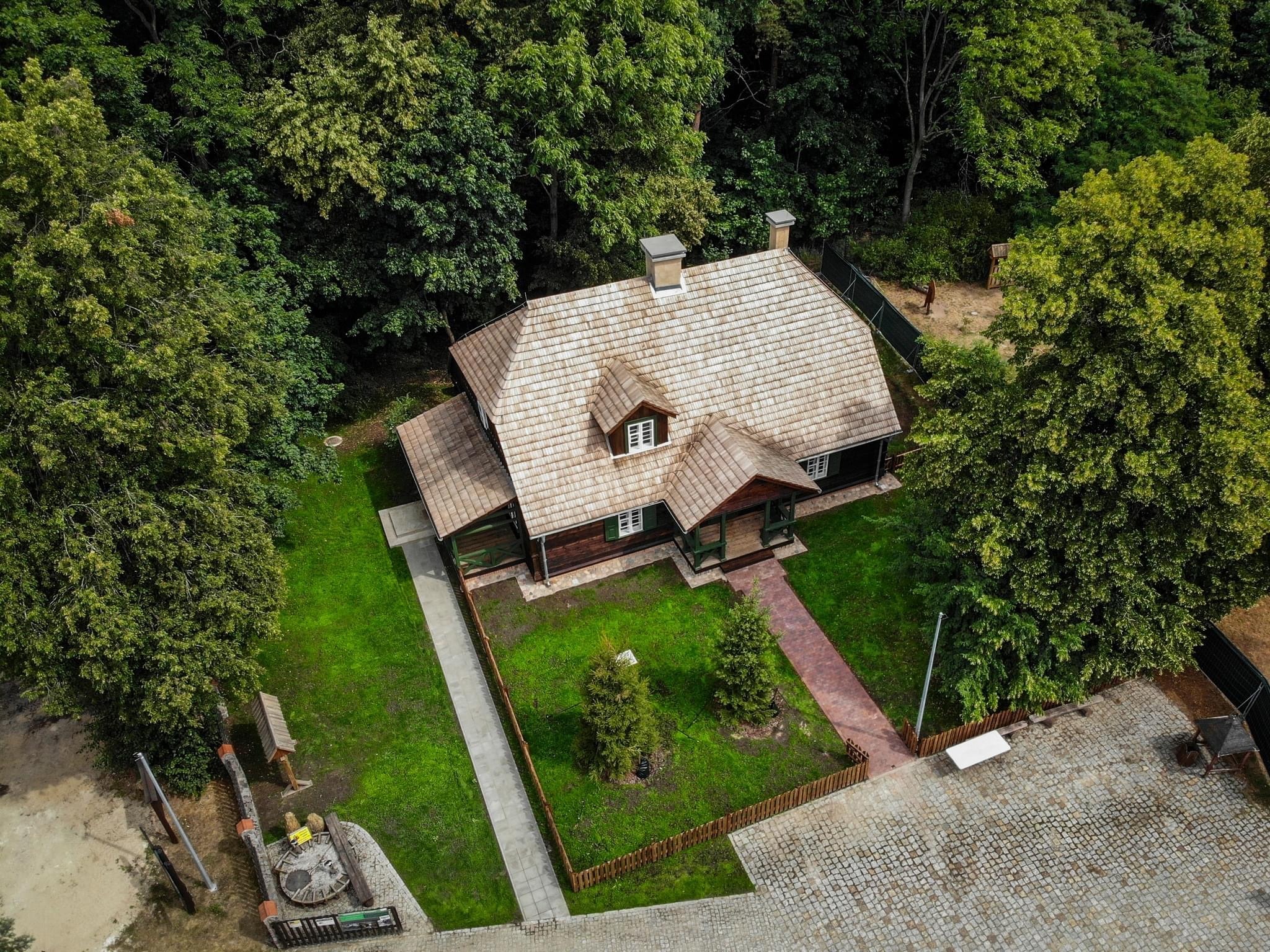 Drewniany dom z dwuspadowym dachem pokrytym gontem, widok z lotu ptaka, otoczony zielenią drzew i trawników, z kamiennymi ścieżkami i placem z kostki brukowej.